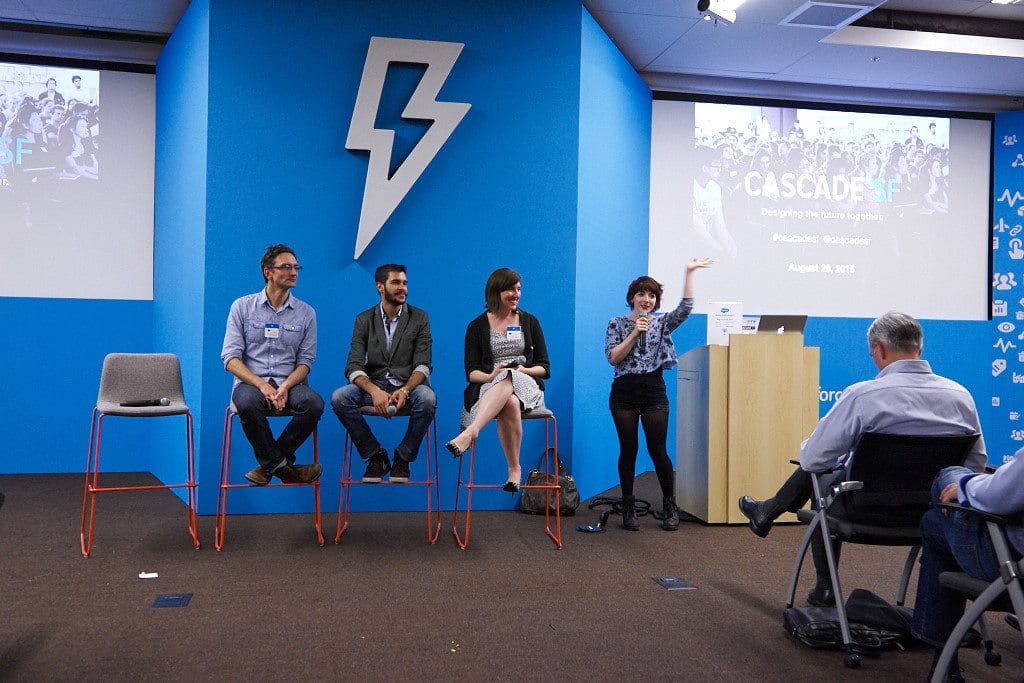 Three panelists seated on stools while Andi Galpern holds a microphone with her arm raised in front of a blue Cascade SF lightning-bolt wall